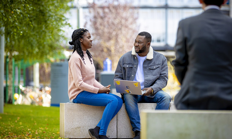 A black man and woman sat outside, chatting together on the university campus.