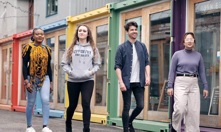 Four students walking through Fargo Village in Coventry, with bright shipping container shops behind them.