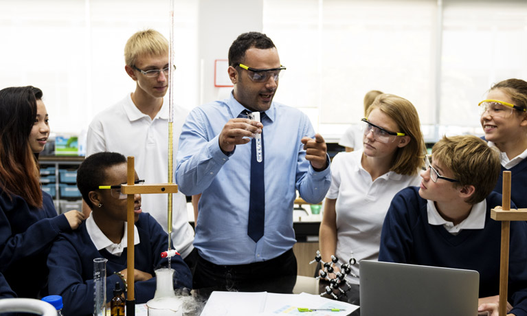 A teacher holding up a test tube in a chemistry lab while students gather round to watch him.