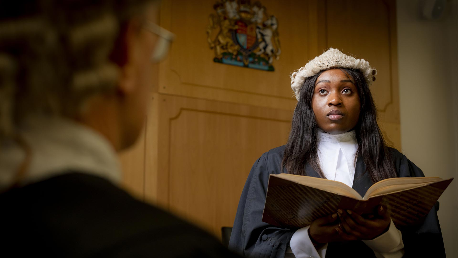 Female student standing in a mock court room wearing  robes and wig, holding an open book