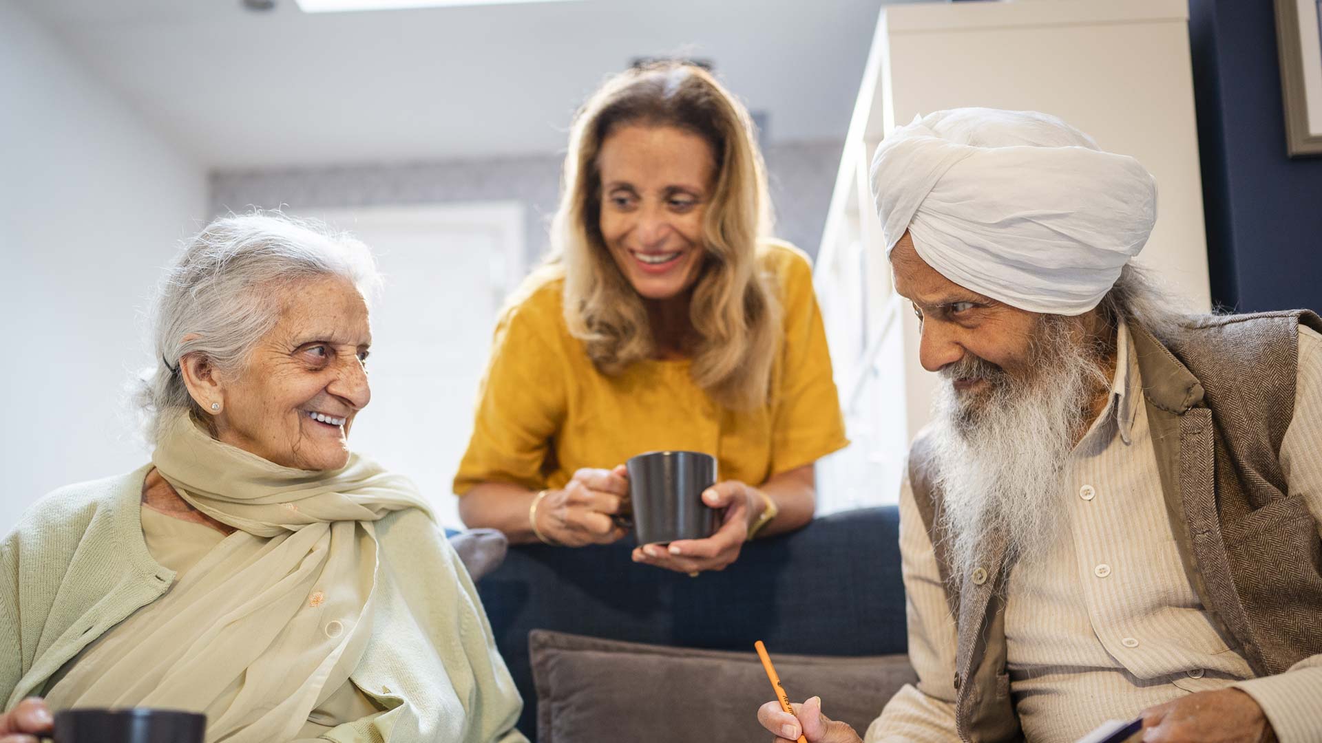 Two elderly people sitting on a sofa drinking tea with a healthcare worker.