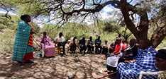 A group of people sat outside in a semi-circle shaded by trees in Africa