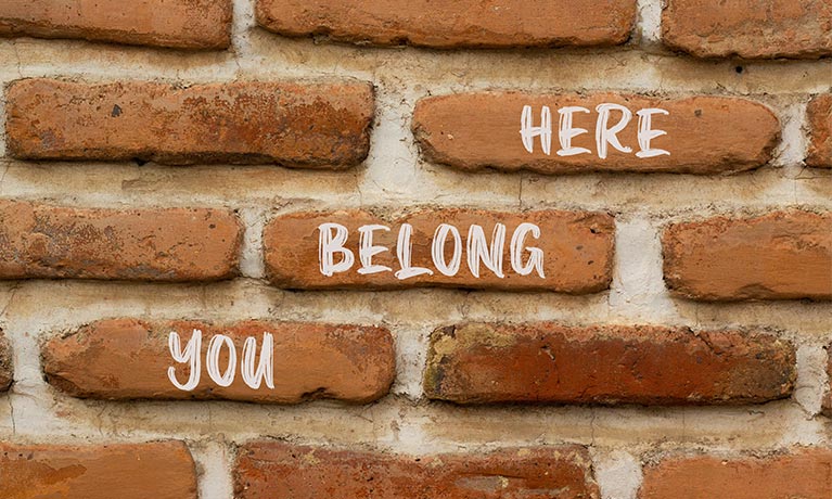 white chalk diagonally reading 'Here you belong' on a brick wall 