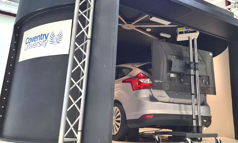 The photo shows a car inside a driving simulator. The car is silver and the simulator exterior is black with a Coventry University logo
