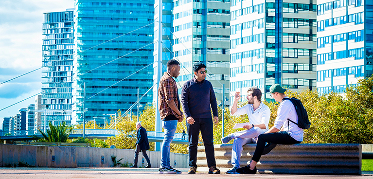 group of students chatting sitting outside on a wall with high rise buildings in the background 