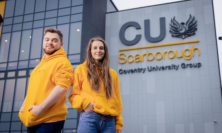 Two students wearing yellow hoodies standing infront of CU Scarborough building