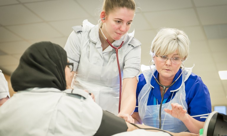 Young nurse and older nursing looking at monitoring equipment whilst taking the blood pressure of a trainee nurse