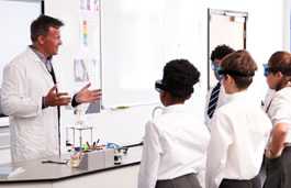 A teacher in a lab coat with a flask on the desk in front of him, talking to a group of students wearing safety glasses.