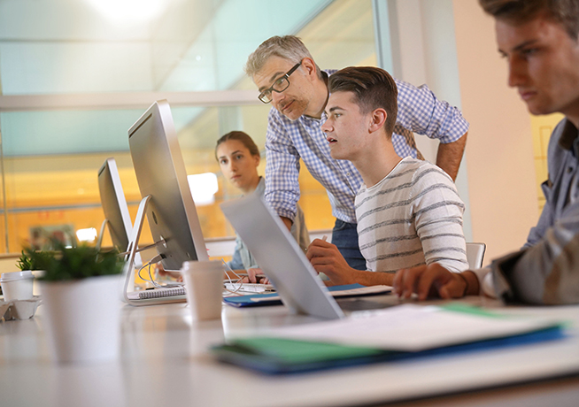 Row of students working on their laptops with a lecturer standing over a student