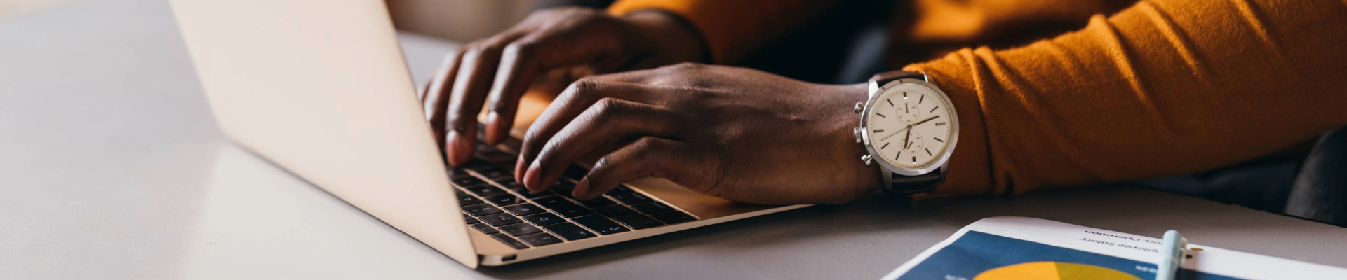 Person typing on a laptop at a desk with a watch on wrist and a printed chart nearby.