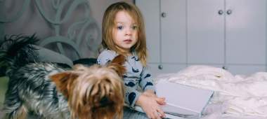 Child sat on a bed holding a book open with a dog