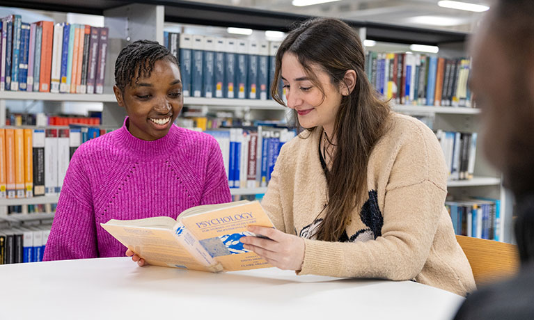 2 female students reading a book together in a library