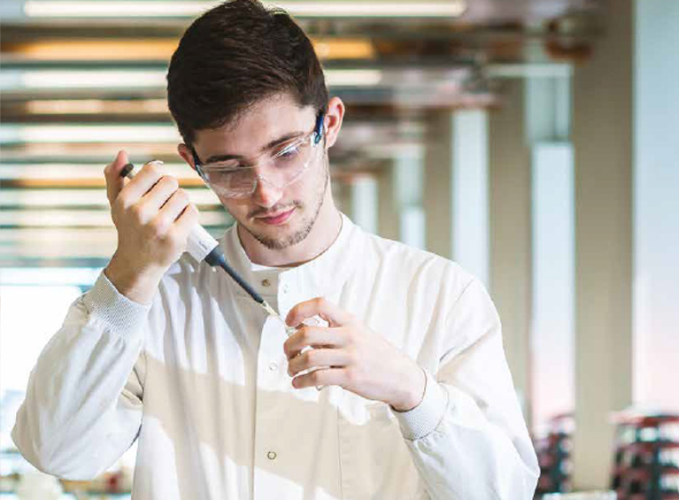 A student in a lab coat using a pipette to dropper liquid into a test tube.