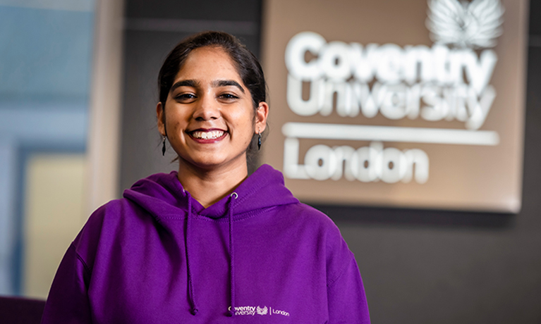 Student ambassador in purple hoody standing in front of 'Coventry University London' sign