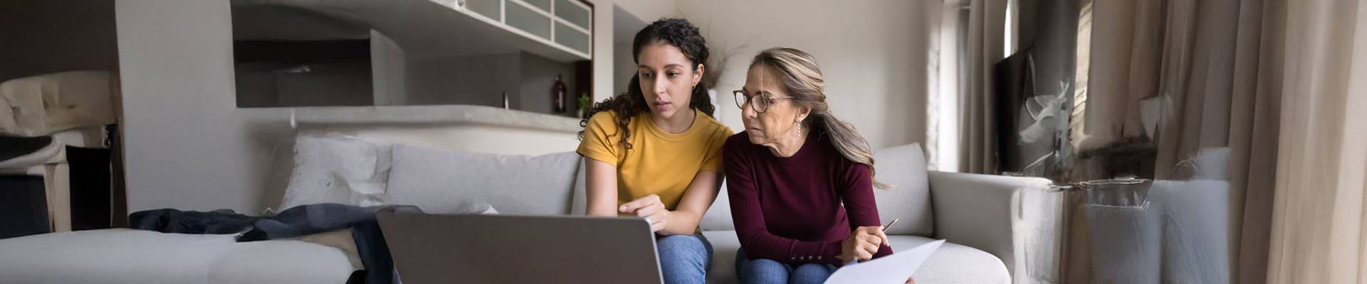Focused Latin young daughter helping senior mother, showing how to use online financial e-bank application on laptop, paying bills on Internet, checking domestic expenses, budget, fees