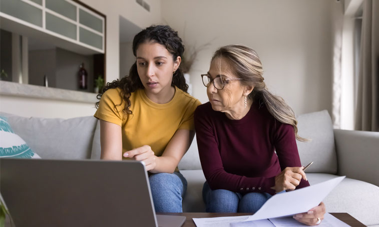 Focused Latin young daughter helping senior mother, showing how to use online financial e-bank application on laptop, paying bills on Internet, checking domestic expenses, budget, fees