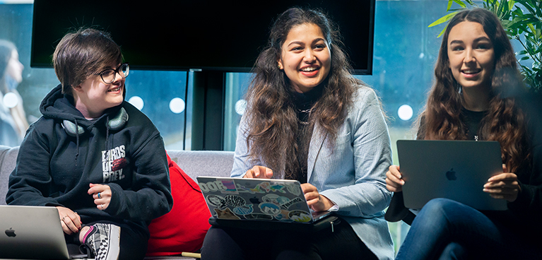 Three students sat on campus smiling with their laptops resting on their laps