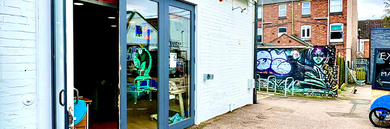 Outside of FabLab Coventry in FarGo Village showing a low building with the FabLab logo on a large glass door next to a wall with graffiti art