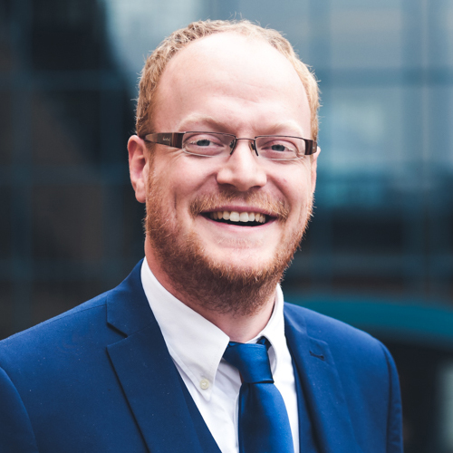 James Malcolm, a smilling man with auburn hair and a short beard wearing glasses and a blue suit and tie
