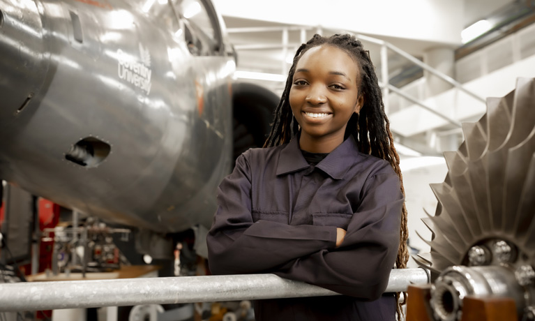 A young woman smiling with folded arms next to a Harrier Jet in Coventry University's engineering building.