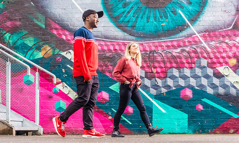 Two students walking together side by side with a brightly coloured wall art besides them