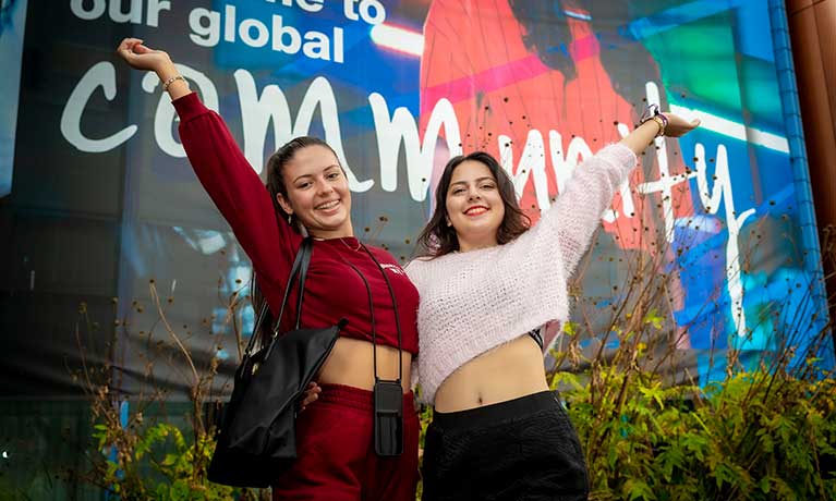 Two female students smiling with hands in the air standing in front of a global community banner 