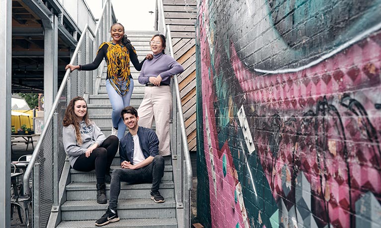 Four students stood on steps outside next a wall of art