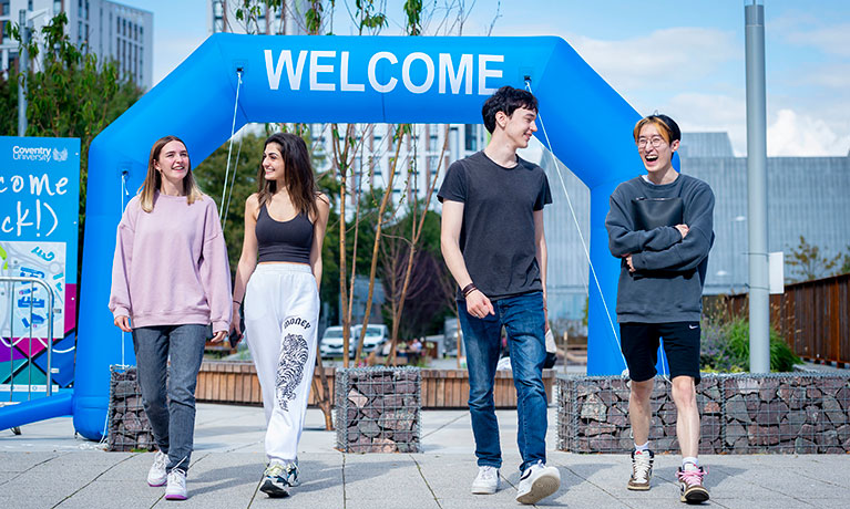 3 females and one male walking on campus with a welcome banner in the background 