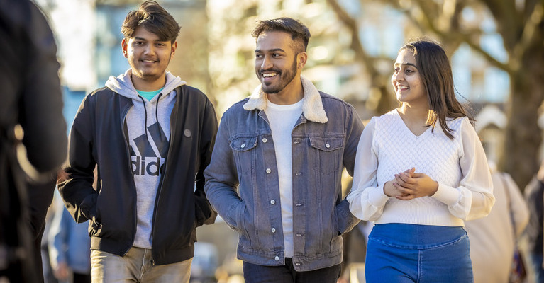 Three students walking by the Thames smiling at the camera