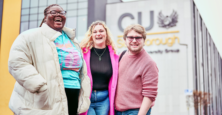 Three students smiling at the camera outside the CU Scarborough campus building.