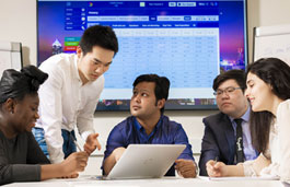 5 students talking at a table around a laptop, with big screen in background.