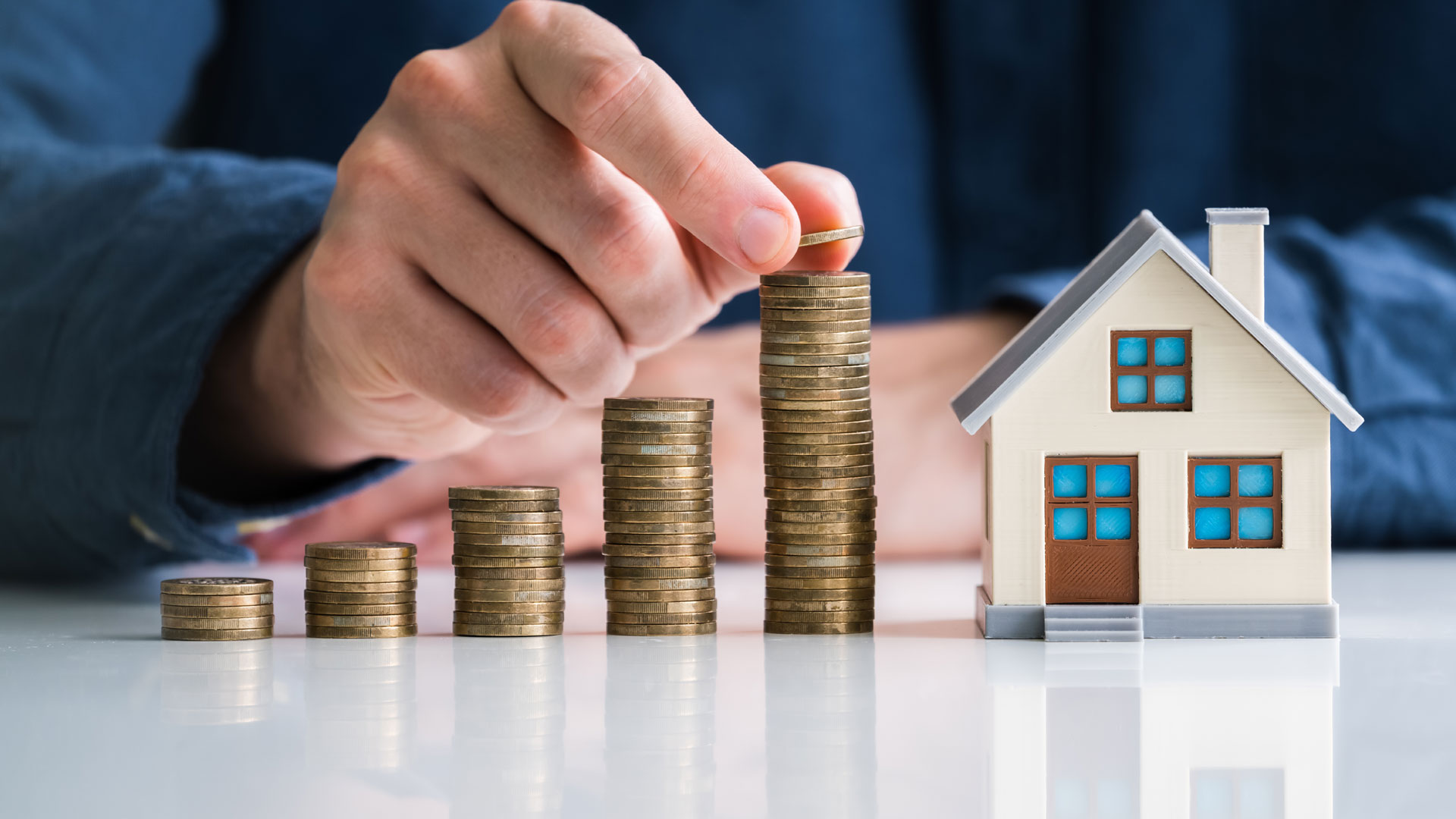 Five stacks of coins with a hand reaching for the highest stack, next to a model house same height as tallest stack