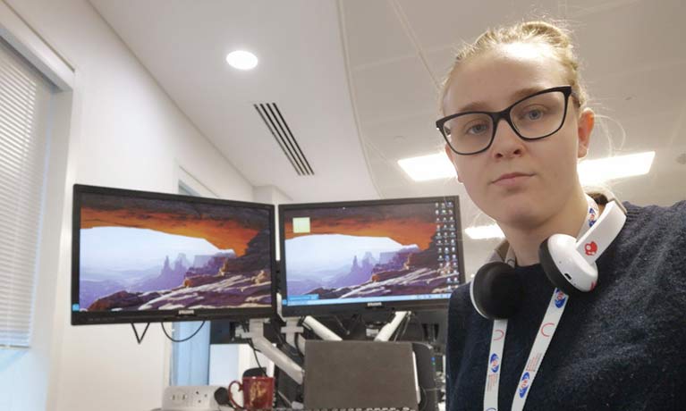 student louise ayres wearing headphones round her neck with 2 screens in the background