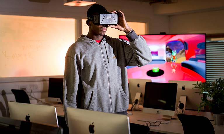 Male student wearing VR glasses in room with mac computers on desks around him