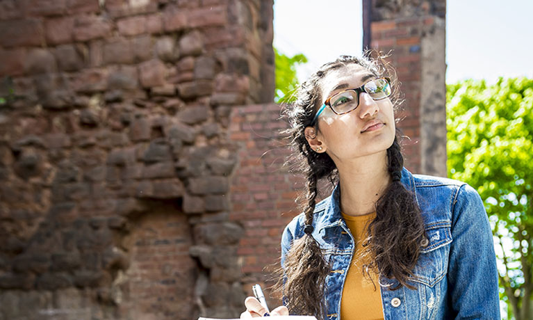 A student outside peering upwards and taking notes with an old brick wall in the background