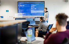 Student stands in focus in background with large screen showing code, two other students on laptop sit between him and the camera