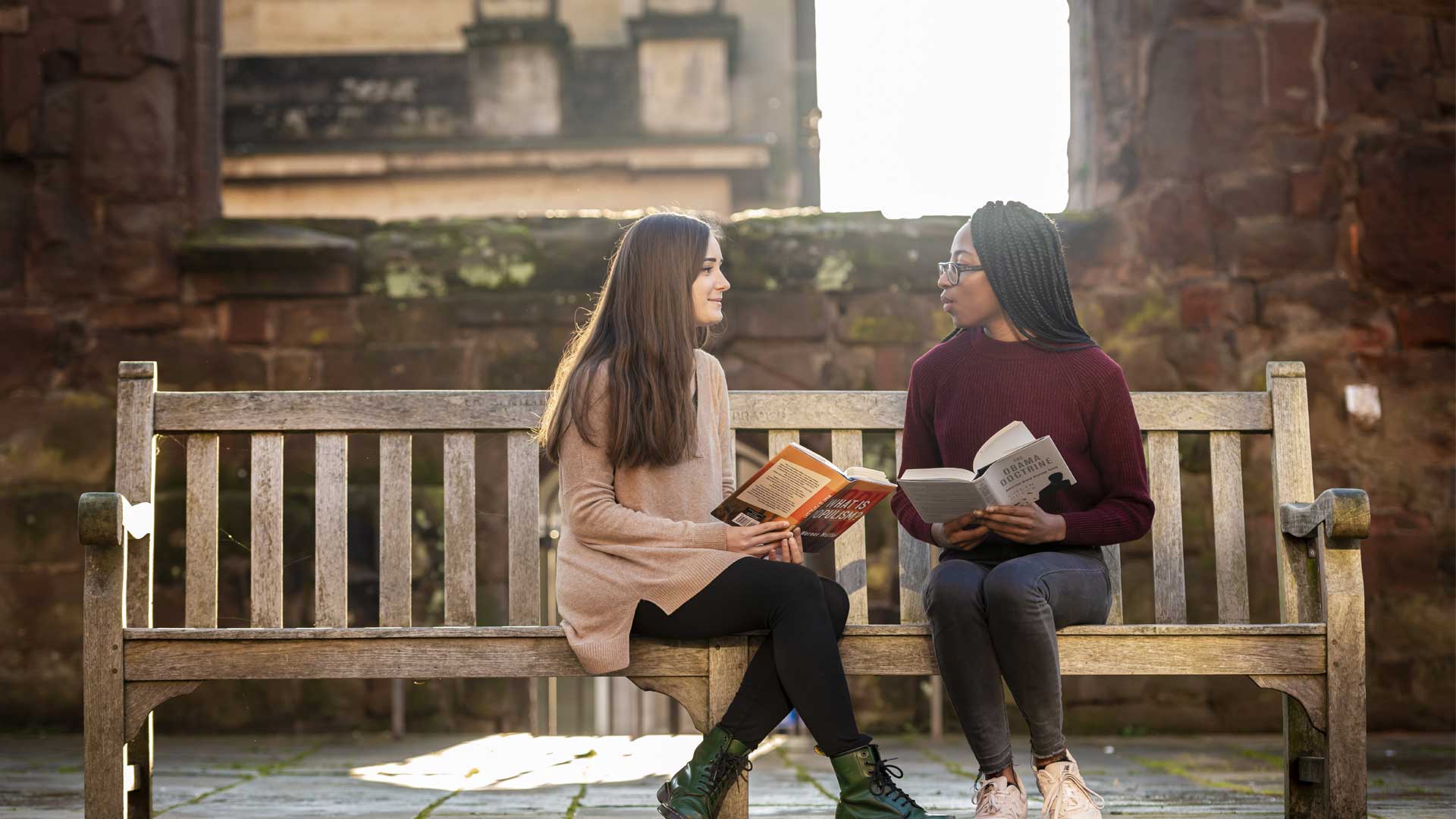 Two students sitting on a bench in front of an old building holding books