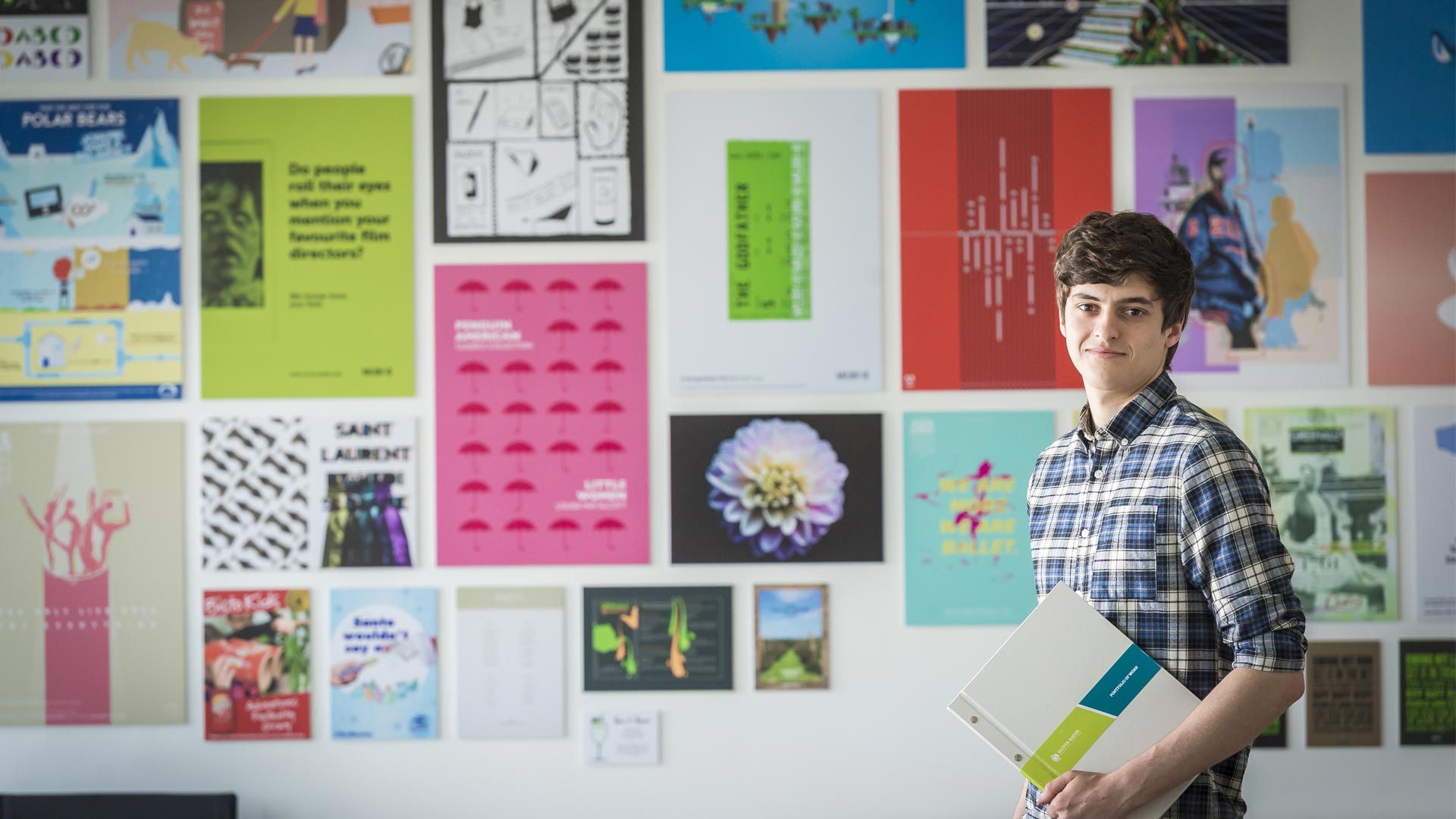 Student holding their portfolio in front of a wall of students' graphic design works 