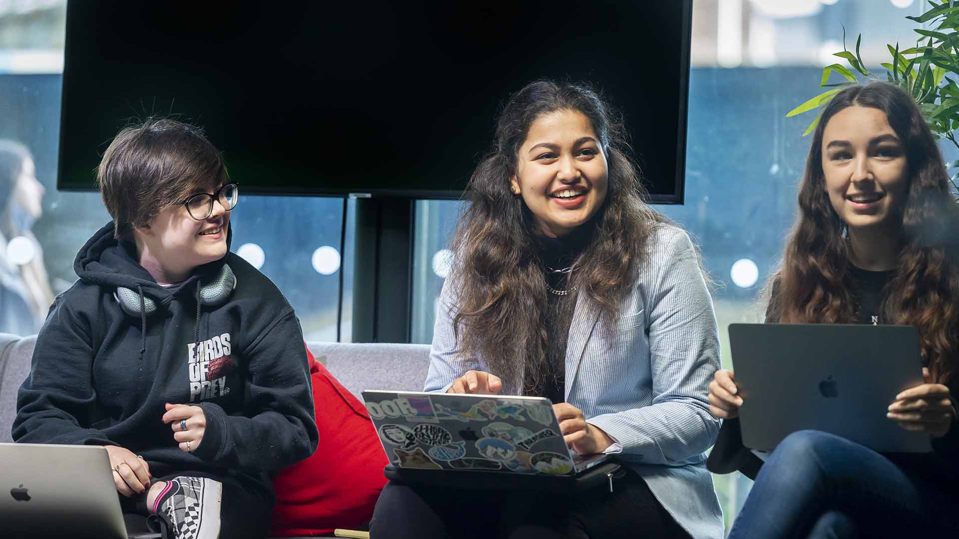 Three students sitting in front of a big screen tv with macbooks