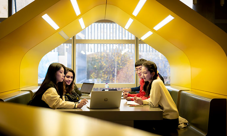 Four people sat in a pod looking at a laptop and talking