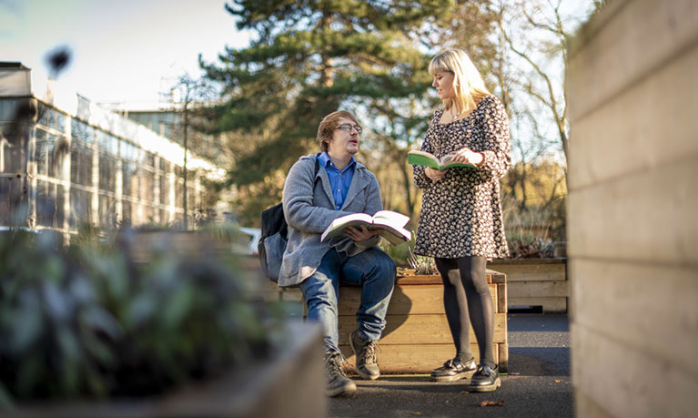 Male student sitting outside with a book in his lap, talking to a standing female student