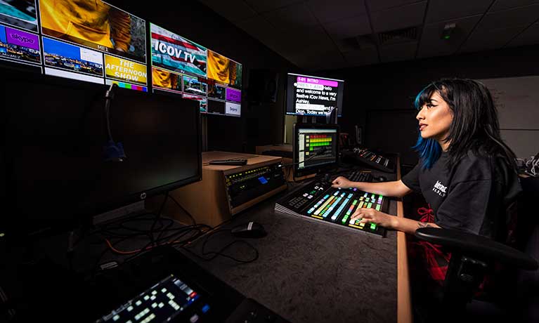 A student working at a computer with multiple screens in a dark lit studio