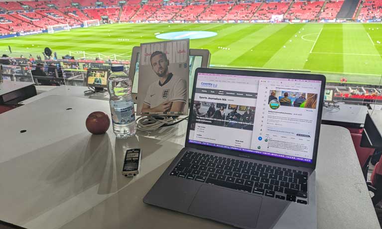 A laptop showing the Sports Journalism page, on a desk with a football stadium in the background