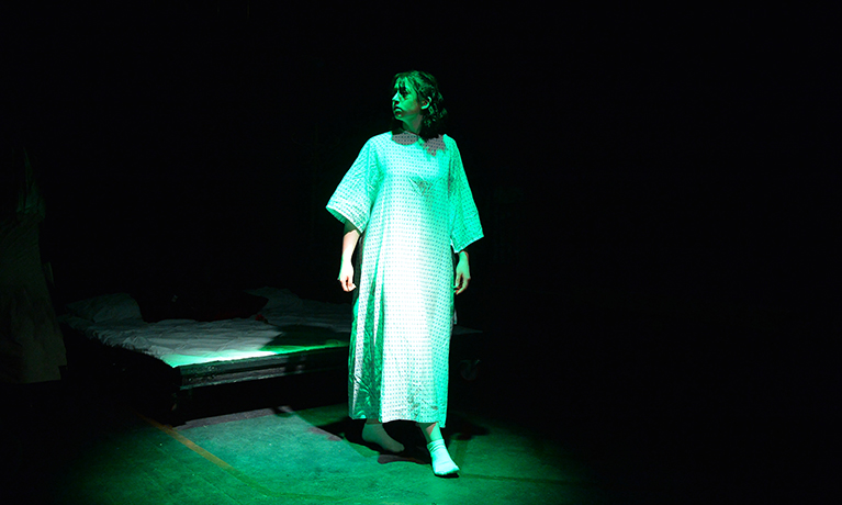 A student acting in a play wearing a white gown and a green spotlight shinning on her