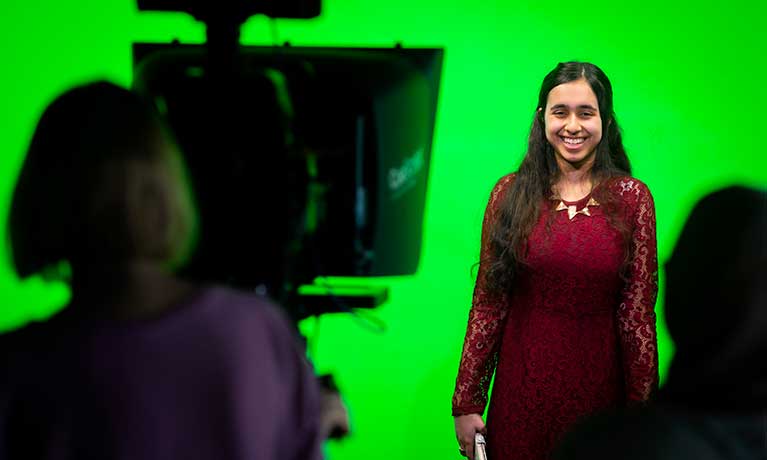 A student stood in front of a film camera with a green screen behind