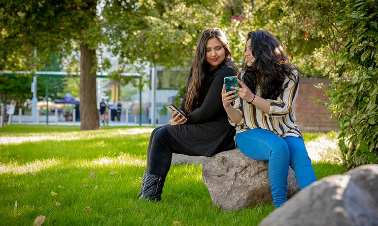 Two students sat in down outside talking and looking at a phone