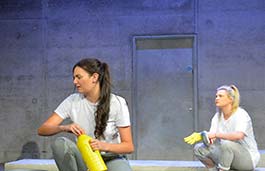 Two students acting on stage sat on the floor with a concrete wall behind and stage lighting