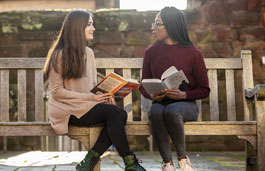 Two students sitting on a bench in front of an old building holding books