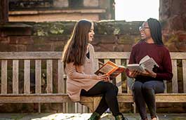 Two females sitting on a bench in front of an old building holding books