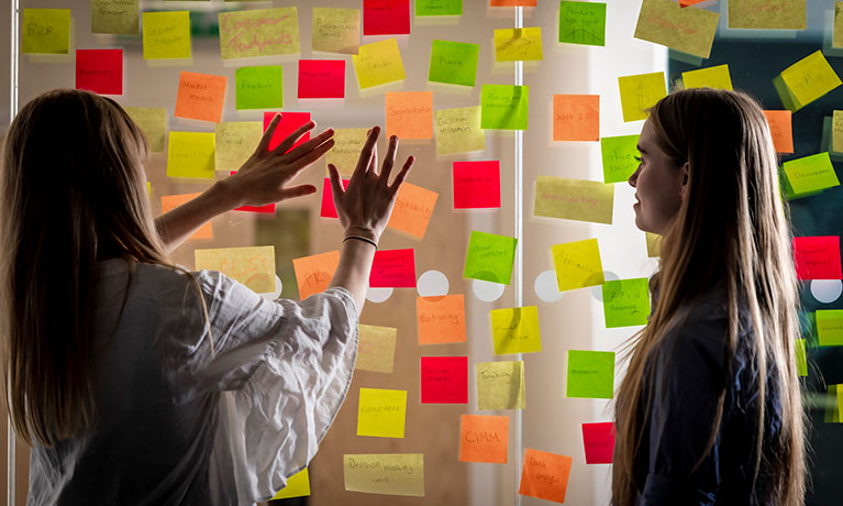 Two young women looking at a glass wall covered in multi-coloured post-its